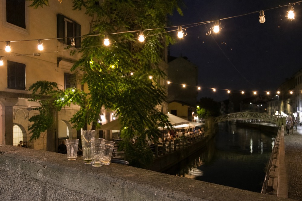 A row of vintage bar signs and string lights above a Navigli towpath with parked bicycles