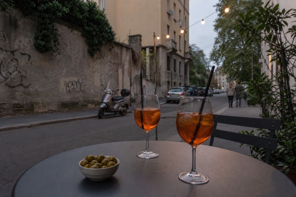 A small round table with a spritz, a plate of olives and a neon sign above a Nolo side street