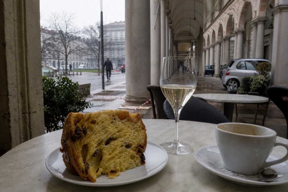 A slice of panettone on a small plate beside a glass of pale sparkling wine and a cup of milky coffee