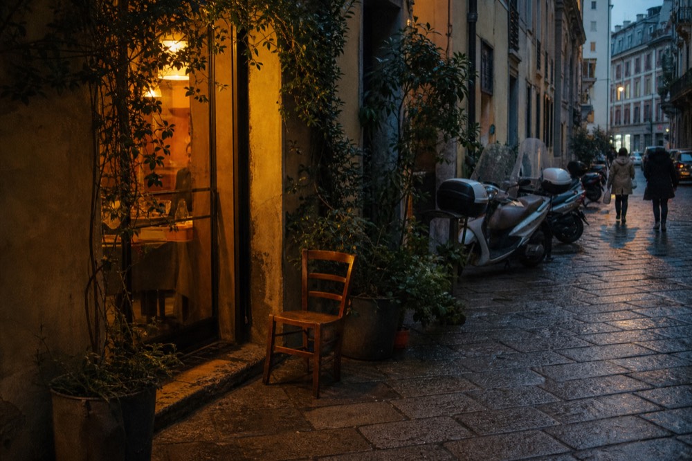 A narrow Milan side street at dusk with a small warmly lit restaurant entrance and bicycles parked outside