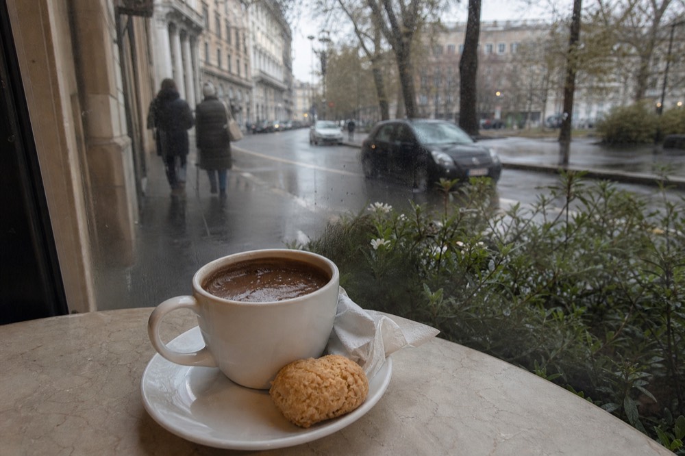 A warm cup of coffee and chocolate drink beside a small pastry on a café saucer
