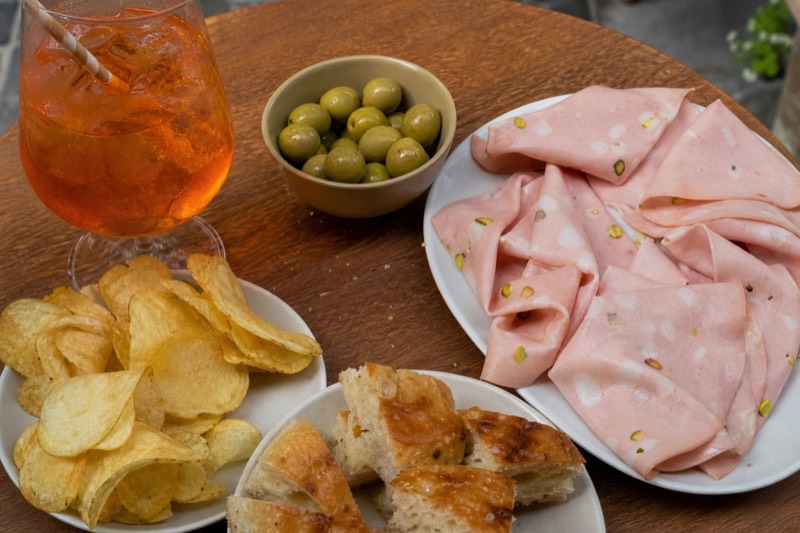An aperitivo table seen from above: an orange spritz, a bowl of green olives, crisps, squares of focaccia and a plate of pistachio-studded mortadella