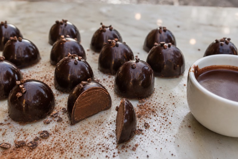 Rows of dark chocolate praline domes dusted with cocoa on a marble counter, one cut open to show the smooth gianduja centre, beside a cup of thick hot chocolate