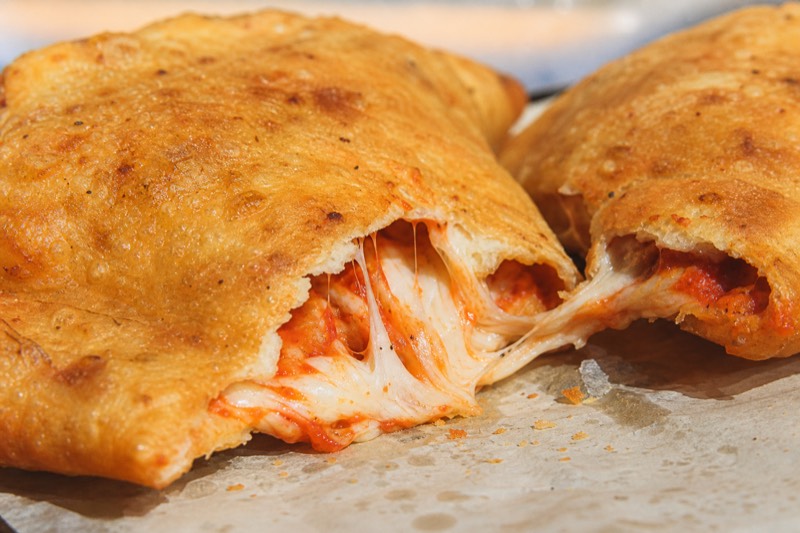 Two golden fried panzerotti on greaseproof paper, one torn open so the melted mozzarella stretches away from the tomato filling