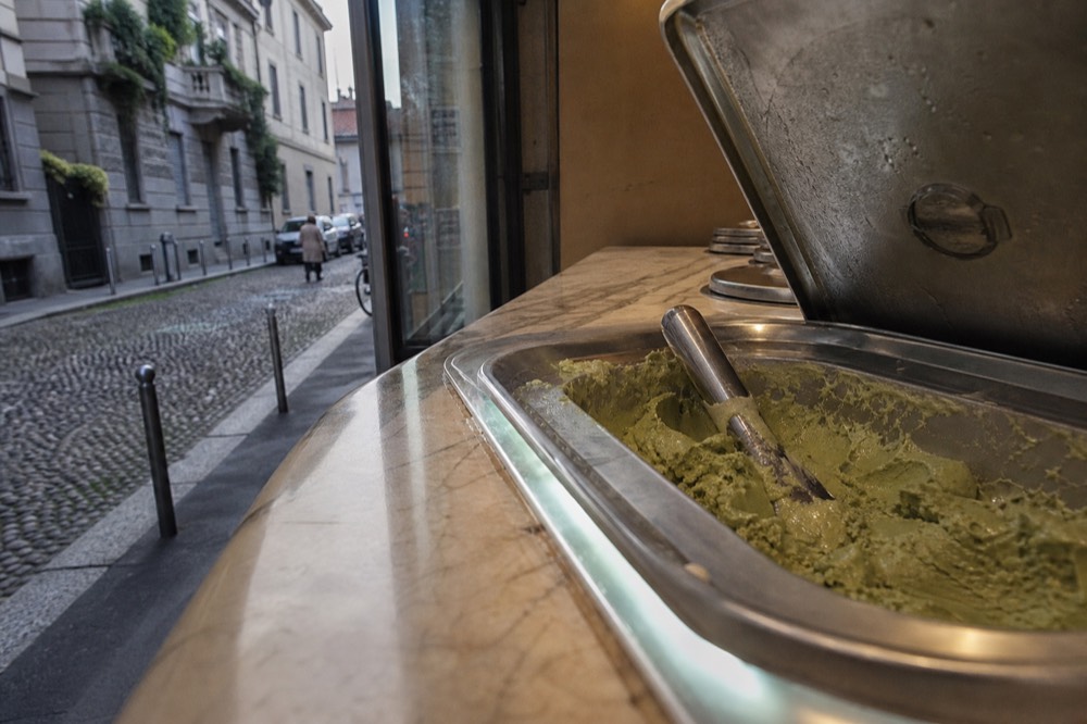 Close view of a stainless steel gelato container with its lid lifted and a metal scoop resting inside