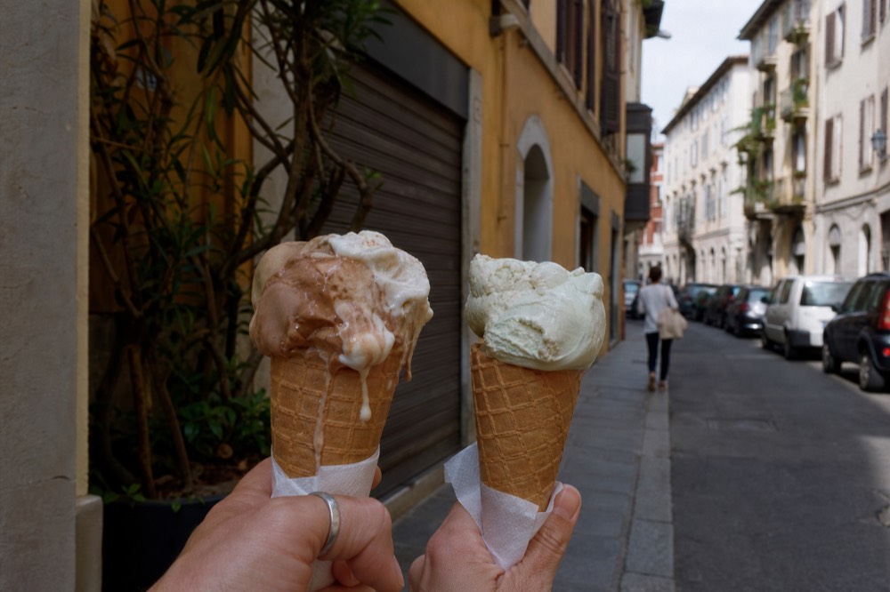 Two gelato cones held above a Milan street with shuttered shopfronts behind