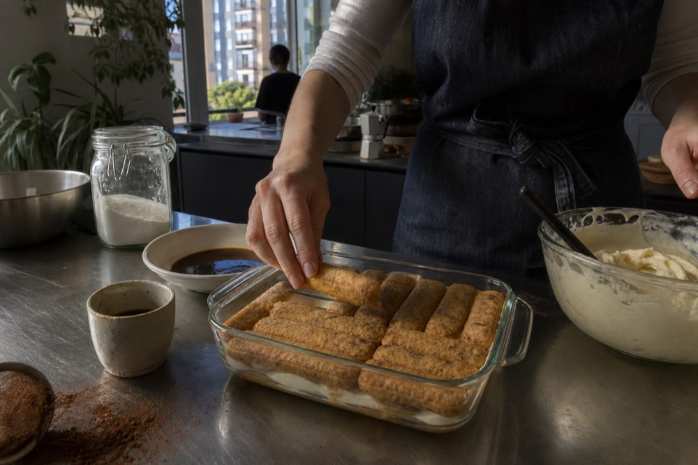A cook layering coffee-soaked sponge into a glass dish for tiramisu