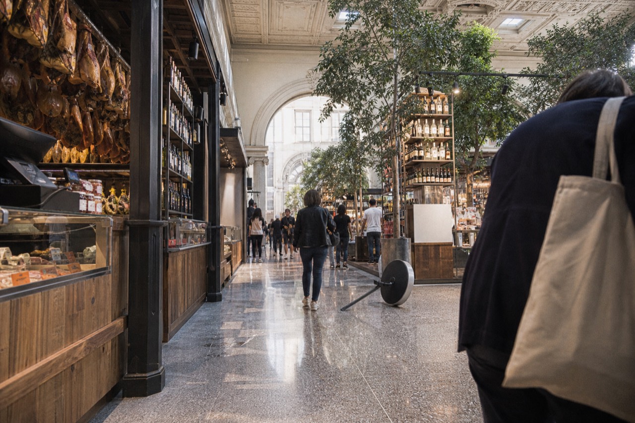 Interior of a large Italian food hall with wooden counters, hanging cured meats and shelves of bottled goods under theatre-style ceilings