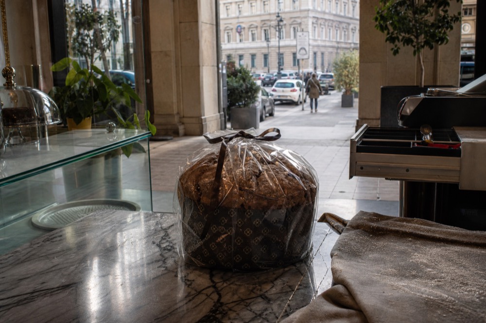 A wrapped artisan panettone with a paper collar standing on a marble pastry shop counter